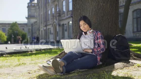 年轻的大学生使用笔记本电脑坐在树下微笑着社交视频的预览图