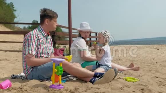 一个穿格子衬衫的成熟男人和两个女孩坐在河边的沙子上家庭户外活动视频的预览图