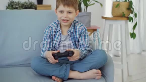 穿着休闲装的快乐小男孩独自坐在家里的沙发上玩电子游戏现代科技快乐视频的预览图