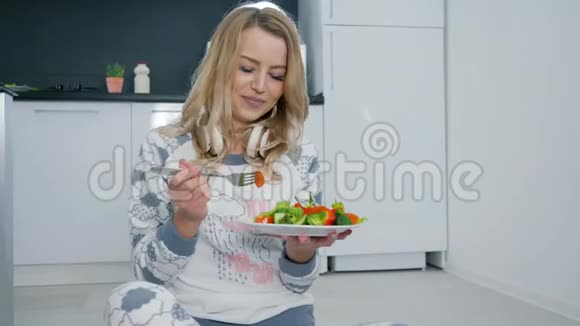 可爱的女孩在厨房里穿着睡衣坐在地板上吃新鲜的健康蔬菜沙拉视频的预览图