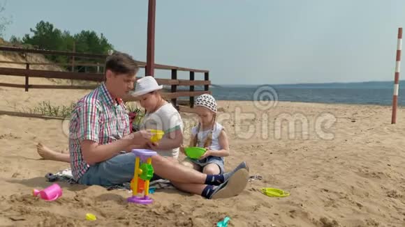 一个穿格子衬衫的成熟男人和两个女孩坐在河边的沙子上家庭户外活动视频的预览图