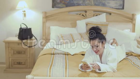 穿着浴袍的漂亮黑发女人躺在酒店房间的床上在电话里交谈视频的预览图