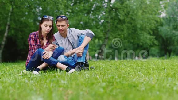 一男一女的朋友坐在公园的草地上享受着智能手机视频的预览图