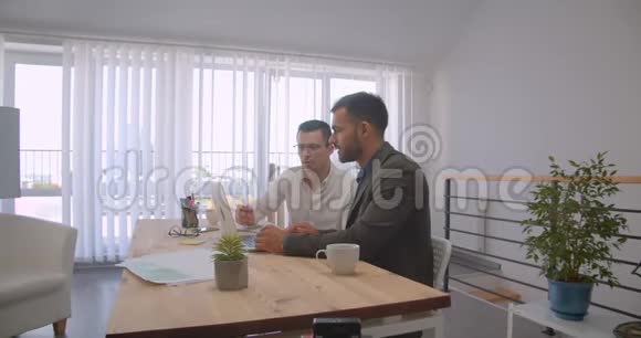 两个成年白种人商人使用笔记本电脑并在办公室内交谈的肖像视频的预览图