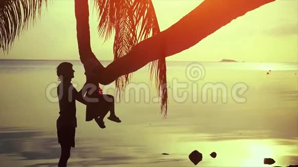 年轻快乐的父亲的剪影在大海的背景下和女儿在棕榈树上玩耍美丽的日落视频的预览图