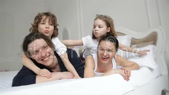 快乐的父母和两个爱笑的孩子正躺在白色的床上在周末的家里一起玩年轻夫妇视频的预览图