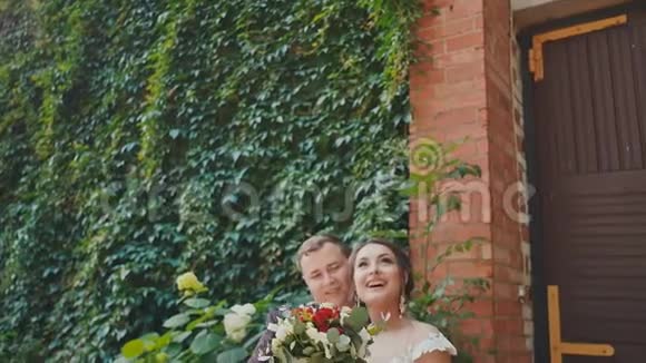 一位优雅的新郎和一位美丽的新娘在砖墙的背景上有一束明亮的花束上面有越来越多的树枝视频的预览图