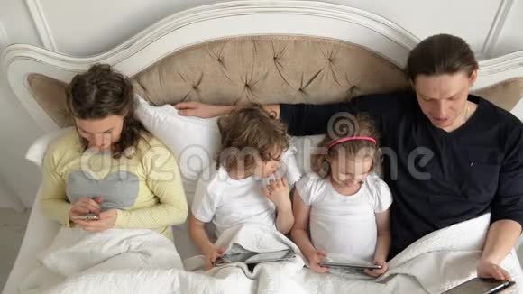 家庭正在使用移动设备躺在床上周末在家两个孩子拿着药片漂亮的妻子视频的预览图