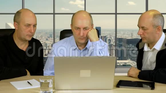 三个商人坐在桌旁拿着笔记本电脑讨论项目拥有全景窗户和摩天大楼的现代化办公室视频的预览图