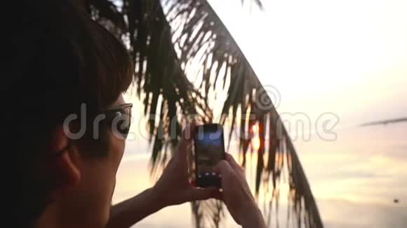 年轻的旅行者戴着太阳镜坐在美丽的夕阳背景下的棕榈树上用一部毫无生气的手机拍照视频的预览图