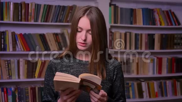 女白种人学生在书架背景上看书和观看镜头视频的预览图