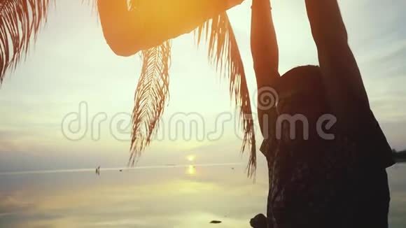 年轻的快乐男子戴着太阳镜挂在棕榈树上在海滩上惊人的日落慢动作1920x1080视频的预览图