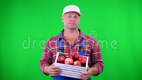 英俊的男农民举着一个木箱里面放着红色成熟的有机苹果微笑着在绿色背景上视频的预览图
