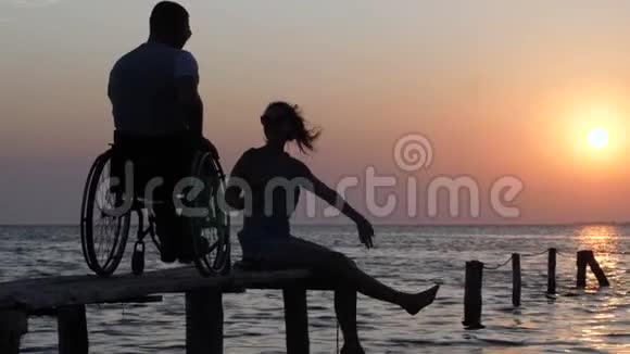 笑的女孩坐在码头上和坐在轮椅上的残疾人在夕阳和大海的背景下交谈视频的预览图