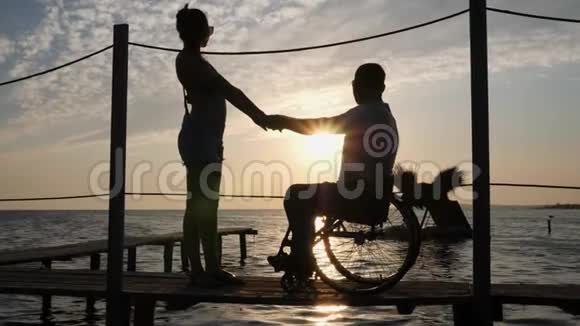 坐在轮椅上的情侣在日落时站在水面和天空的背景上的小剪影视频的预览图