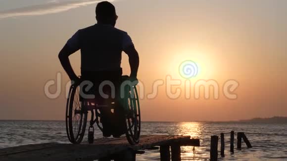坐在轮椅上的残疾人在橙色的天空和夕阳下的水的背景下休息到海港码头视频的预览图