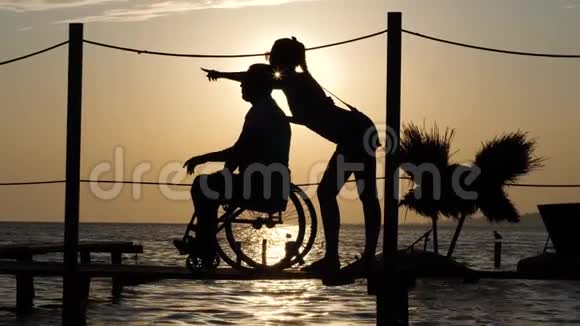 幸运的残疾人和女朋友看着远方在夕阳西下的海面上向地平线伸出双臂视频的预览图