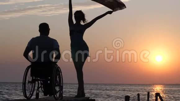 美丽的女孩站在码头上在夕阳的背景下在残疾人面前挥舞着布坐在轮椅上视频的预览图