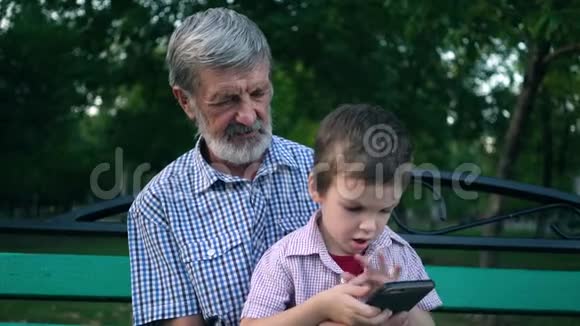 年长的祖父和孙子坐在公园的长凳上玩智能手机视频的预览图