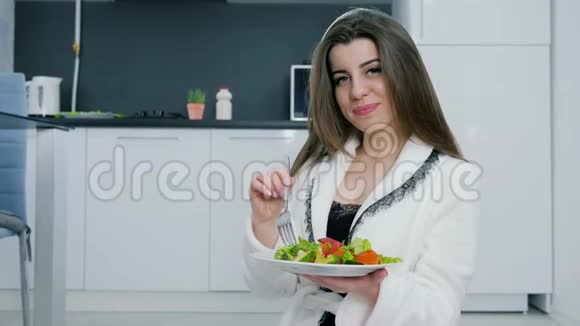 穿白色浴袍的女孩画像手里拿着蔬菜沙拉在厨房背景上视频的预览图