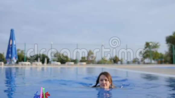 美丽的女性游泳进入蓝色游泳池喝五颜六色的鸡尾酒竖起大拇指视频的预览图