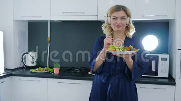 女人戴着耳机听音乐手拿沙拉和叉子在厨房里走来走去视频的预览图