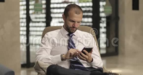 时尚商人使用手机坐在酒店大堂或办公室视频的预览图
