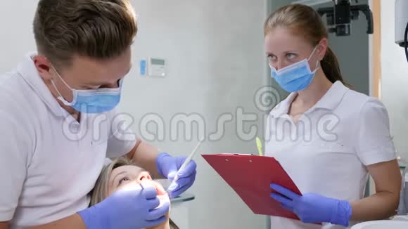 医生用牙科器械抱在怀里看着女孩的嘴旁边的助手把诊断写在记事本上视频的预览图