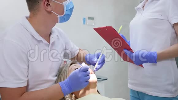 医学检查口腔医生用手拿着牙镜看着女性牙齿旁边的护士写诊断书视频的预览图