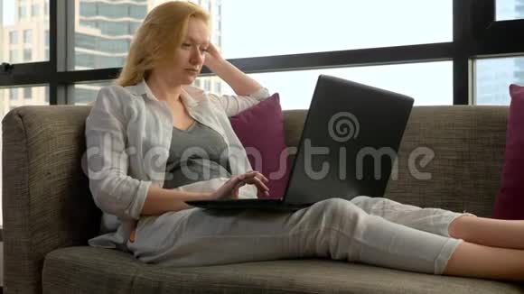 一位优雅的女士躺在沙发上坐在全景窗旁俯瞰摩天大楼并使用她的笔记本电脑视频的预览图