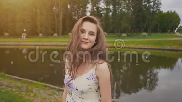 在池塘边的公园里穿着夏装的年轻漂亮女孩女孩微笑着走进镜头把她的头发伸直视频的预览图