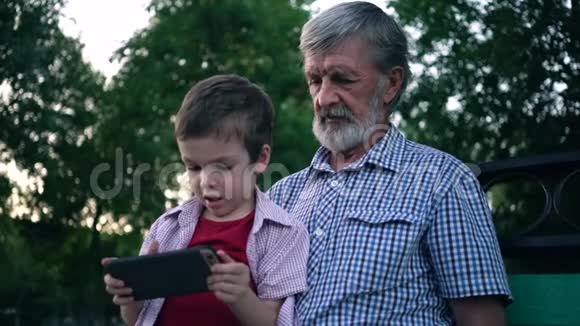 年长的祖父和孙子坐在公园的长凳上玩智能手机视频的预览图