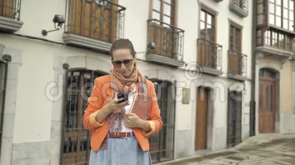 穿着红色夹克蓝色牛仔裤裙子橙色围巾和太阳镜的漂亮女孩用智能手机做面部护理视频的预览图