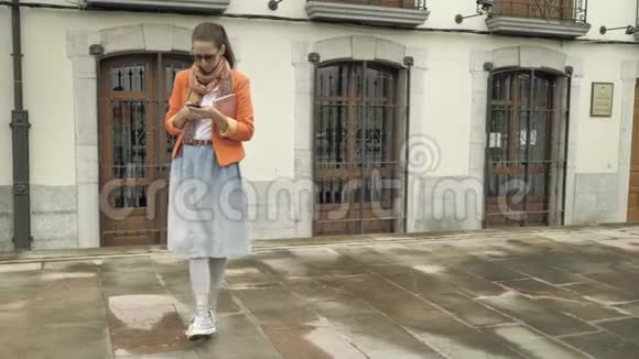 穿着红色夹克蓝色牛仔裤裙子橙色围巾和太阳镜的漂亮女孩用智能手机做面部护理视频的预览图