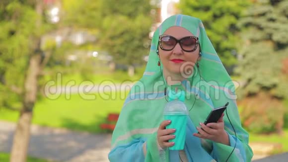 公园里一位戴着浅色围巾的年轻穆斯林妇女正在喝咖啡带着耳机在信使里说话视频的预览图