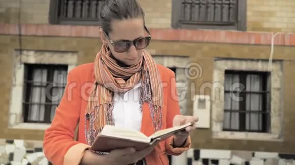 穿着红色夹克蓝色牛仔裤裙子橙色围巾和太阳镜看书的漂亮女孩视频的预览图