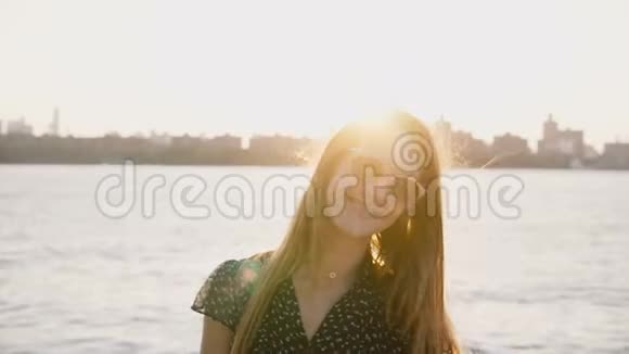 快乐轻松的白种人女孩的肖像戴着太阳镜在令人惊叹的城市天际线海滩日落4K的镜头前微笑视频的预览图