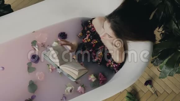 上景一位美丽的年轻女子读了一本书在充满牛奶和芬芳花蕾的温水浴中放松视频的预览图