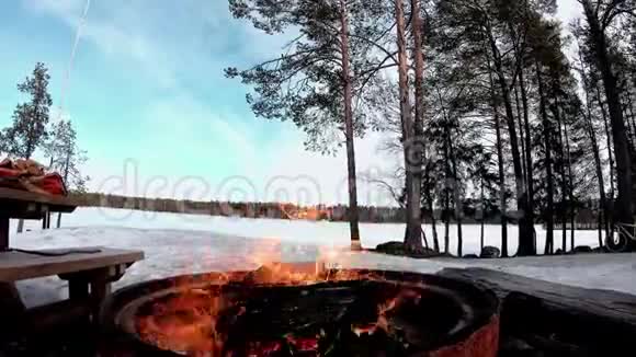 在阳光明媚的日子里用木头烧烤火在白雪覆盖着树木的土地上在美丽的风景有着冻结的湖泊视频的预览图