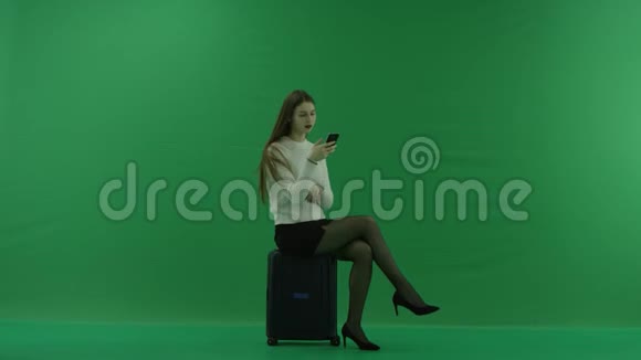 女孩坐在一个手提箱上半转看着右边在绿色屏幕上用手机写一些东西视频的预览图