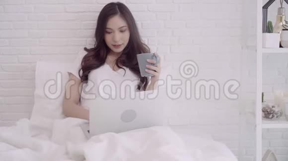 亚洲妇女使用电脑或笔记本电脑拿着一杯温暖的咖啡或茶躺在床上当她在家里的卧室放松视频的预览图