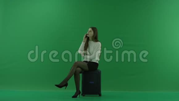 女孩坐在一个手提箱上半转看着左边在绿色屏幕上通过电话交谈视频的预览图
