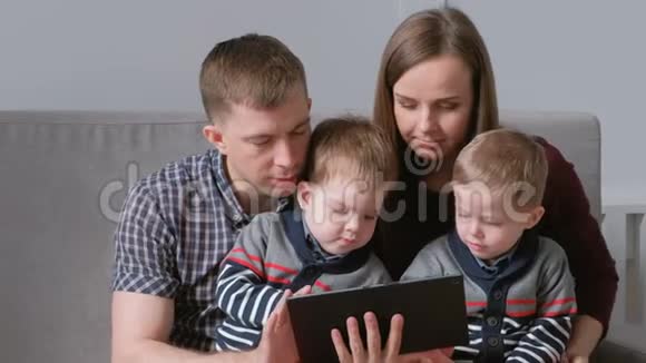 带平板电脑的家人妈妈爸爸和两个儿子双胞胎看着坐在沙发上的平板电脑视频的预览图