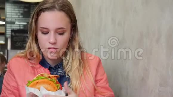 一个吃汉堡包的年轻女孩坐在咖啡馆的桌子旁视频的预览图