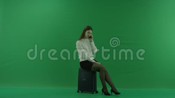 女孩坐在一个手提箱上半转看着右边在绿色屏幕上通过电话说话视频的预览图