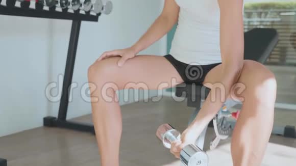 年轻女子坐在长凳上在体育俱乐部用哑铃做二头肌锻炼视频的预览图