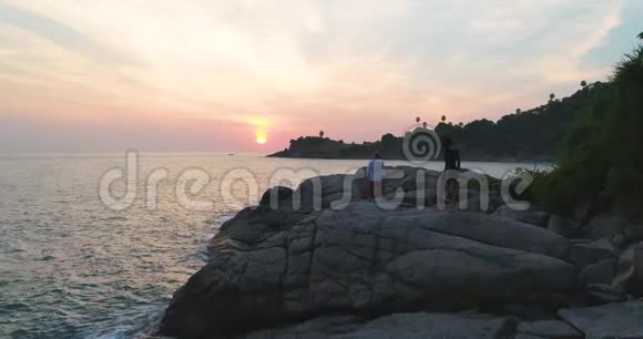 空中一对相爱的夫妇在岩石上看着大海上的日落视频的预览图