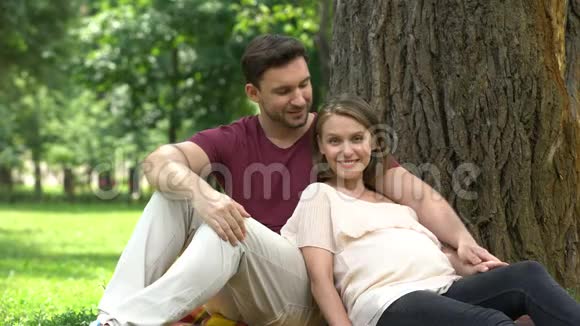 坐在公园里的一对怀孕的夫妇微笑着进入镜头快乐的分娩养育孩子视频的预览图