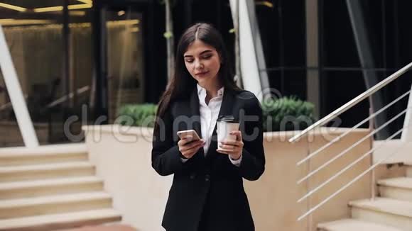 专业的年轻女商人站在城市街道上使用智能手机喝咖啡她看着摄像机视频的预览图