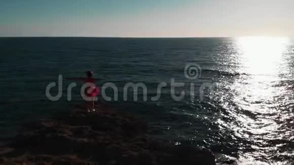 岩石海滩上的女孩站在海崖上的年轻女子穿红色裙子的女孩在海上迷人的女人看日落美丽美丽视频的预览图